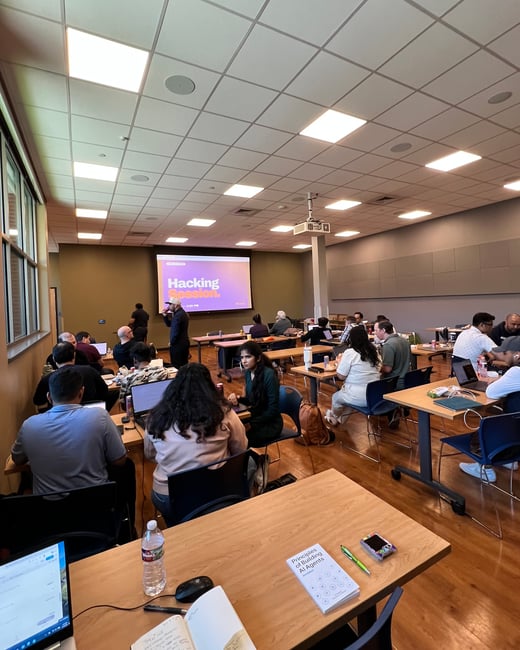 A group of people participating in a 'Hacking Session' workshop in a large room with multiple tables, laptops, and a projector screen.