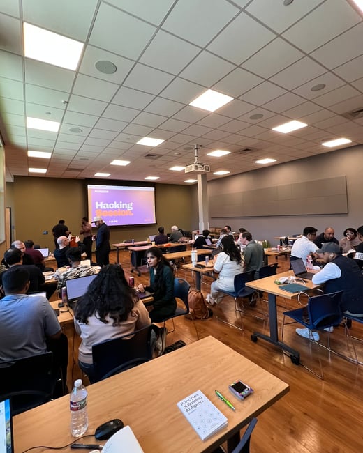 A group of people are participating in a 'Hacking Session' workshop in a classroom-style room, with many working on laptops at wooden tables.