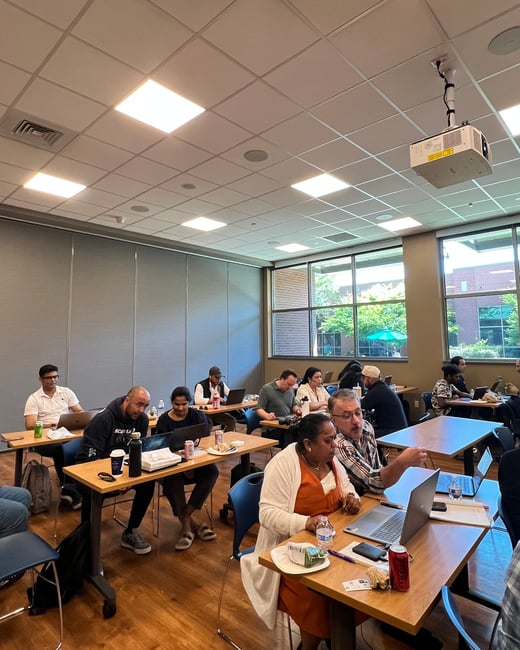 A group of people are gathered in a well-lit room, sitting at wooden tables with laptops open, appearing to be in a workshop or training session.