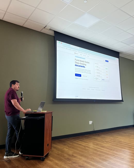 A man in a maroon polo shirt is giving a presentation, standing at a wooden podium with a laptop and holding a microphone. Behind him, a large projection screen displays a website for a service called stip.ai.