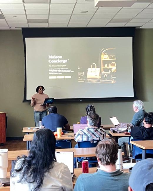 A woman is giving a presentation in a room where several people are seated at tables with laptops, facing a large projector screen.