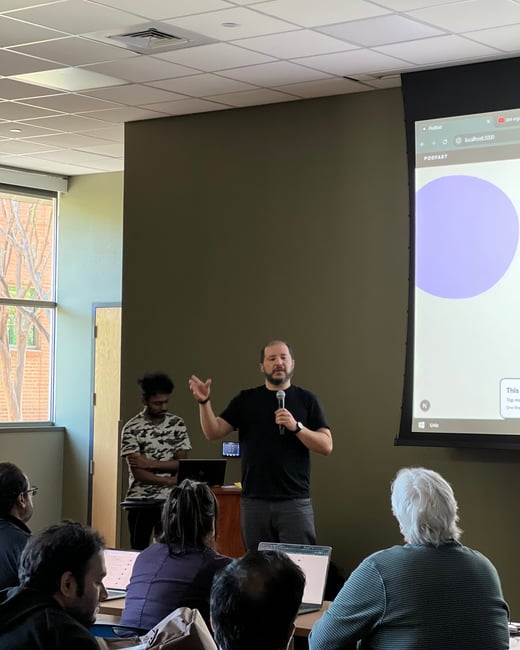 A man is giving a presentation in a room, holding a microphone in front of a projector screen showing a website named 'PODFAST'.