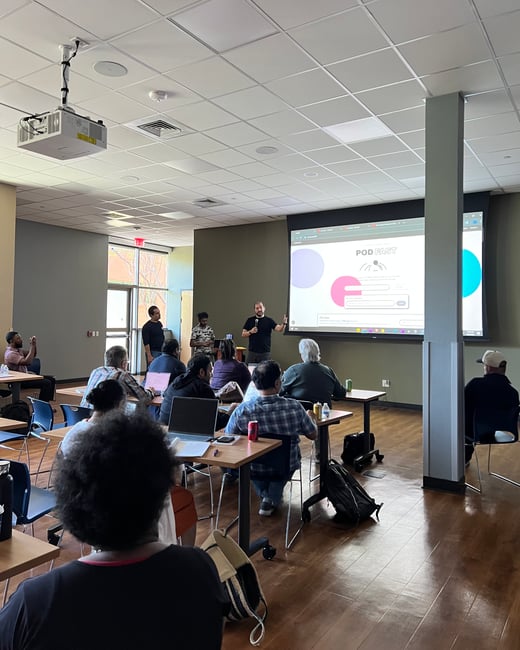 A man is giving a presentation in a classroom or meeting room, with an audience seated at tables and a large projector screen displaying a website titled 'POD FAST'.