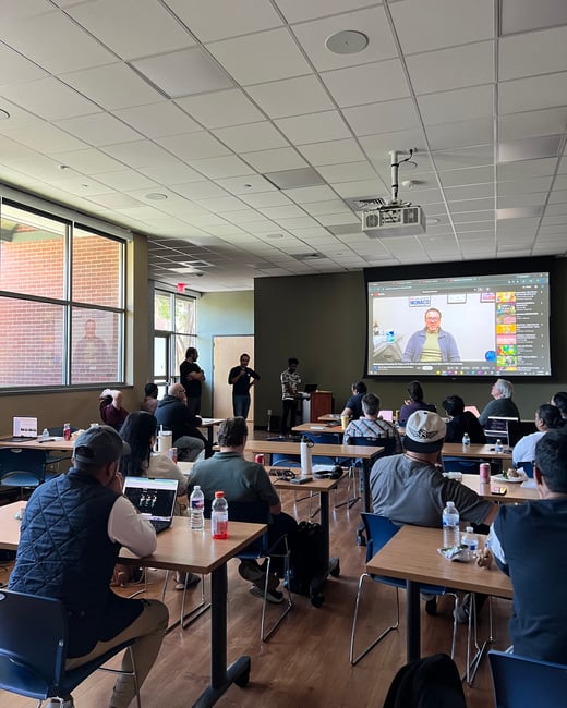 A photograph of a presentation or lecture taking place in a classroom-style room, with attendees seated at tables and speakers at the front near a large screen.