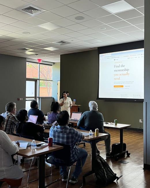 A woman stands at the front of a room giving a presentation to a seated audience, with a large screen displaying a website about mentorship.