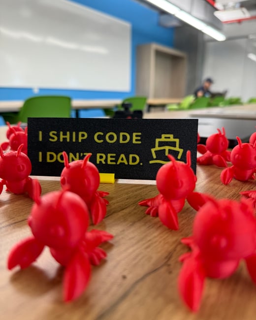 A close-up photograph of several small red 3D-printed crab figurines scattered on a wooden desk, with a black sign in the background that reads 'I SHIP CODE I DON'T READ.'