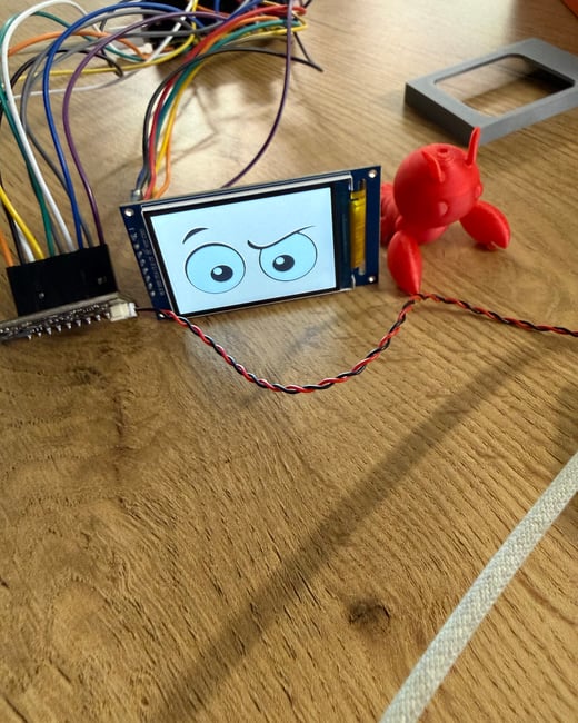 An electronic project featuring an LCD screen displaying skeptical cartoon eyes, connected by a bundle of colorful wires on a wooden surface next to a red 3D-printed figure.