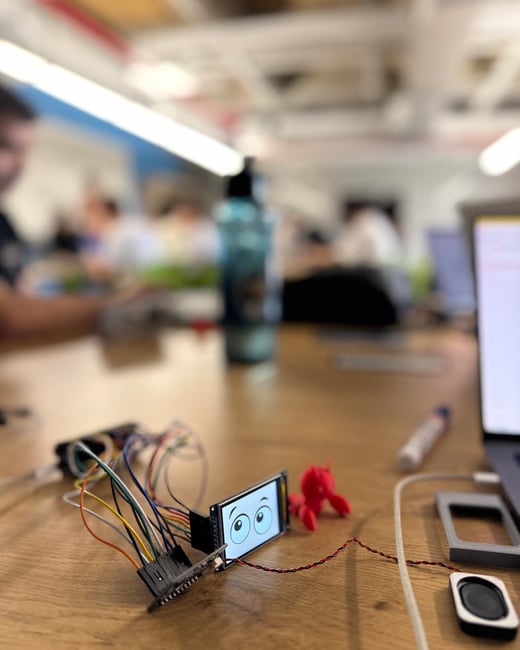 A close-up photo of a small electronic screen displaying cartoon eyes, connected to a circuit board with colorful wires on a wooden desk in a blurred office setting.