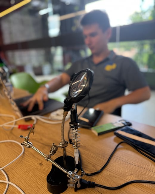 A close-up photograph of a 'helping hand' tool with a magnifying glass on a wooden desk, with a person working on a laptop in the blurred background.