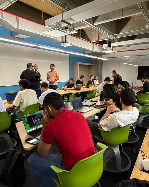 A group of people are engaged in a workshop or coding session in a modern office space, with several individuals working on laptops and others standing near a whiteboard.