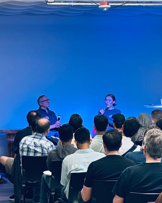 A vertical photograph capturing an indoor event where two speakers, a man and a woman, are seated on a low stage against a brightly lit blue backdrop, addressing an audience seated in rows in the foreground.