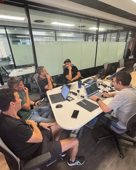 A group of five men are gathered around a white table in a modern office setting, engaged in a collaborative discussion with several open laptops.