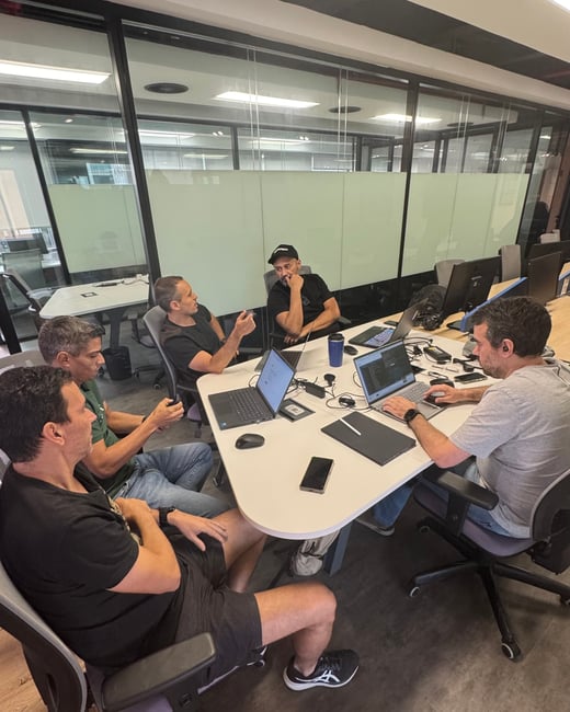 A group of five men are seated around a white table in a modern office, engaged in a discussion with several open laptops and a smartphone on the table.