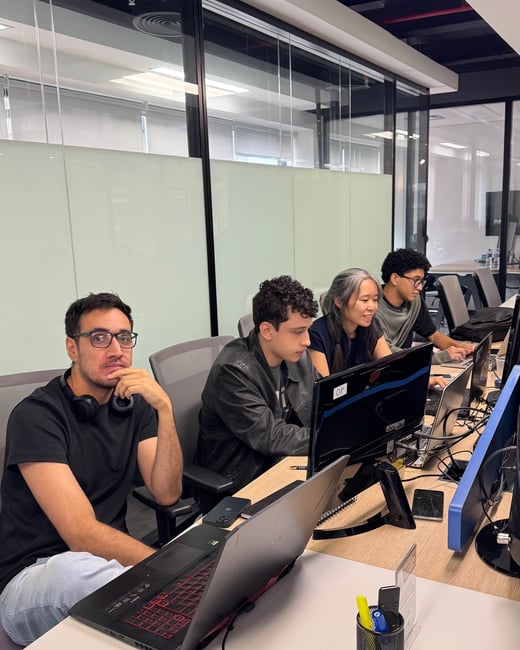 A group of four people are sitting at a long desk in a modern office environment, focused on their laptops and computer monitors.