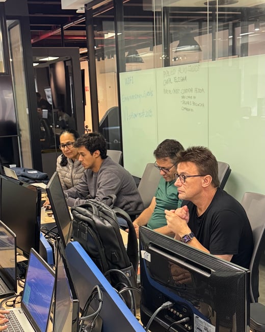 A group of four people are working intently at a long desk filled with computer monitors and laptops in a modern office environment.