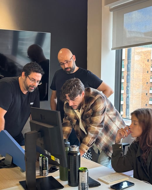 A group of four colleagues are gathered around a computer monitor in an office setting, seemingly engaged in a collaborative task or debugging session.