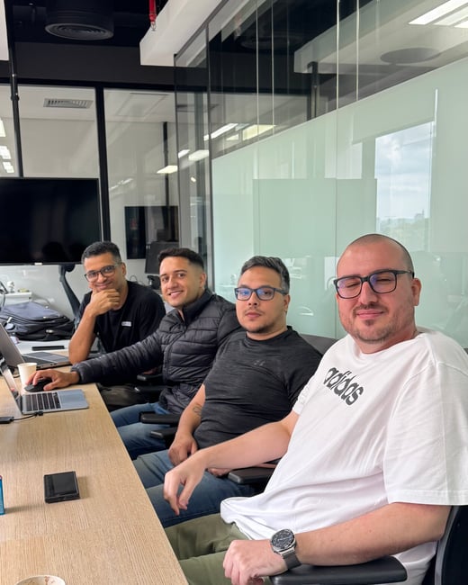 Four men are sitting at a long wooden desk in a modern office setting, all looking towards the camera.