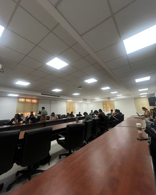 An indoor photograph of a large conference room or classroom where several people are seated at long wooden tables with laptops.