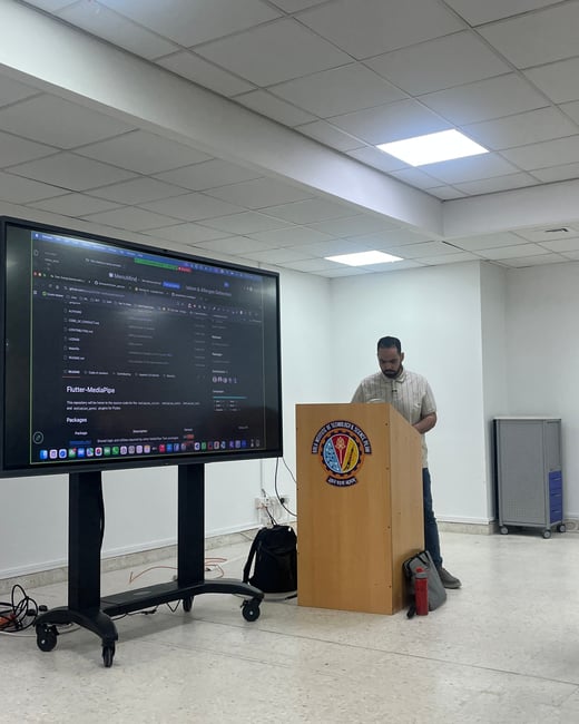 A man stands behind a wooden podium presenting next to a large screen displaying a GitHub repository page.