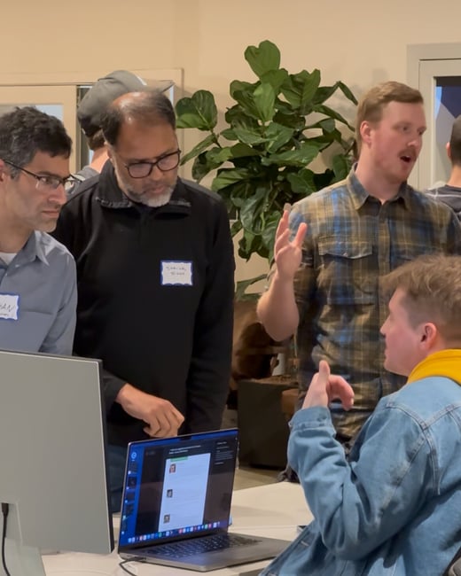 A candid indoor photograph capturing a group of men gathered around a table with a laptop and monitor, engaged in an animated technical discussion or meeting.