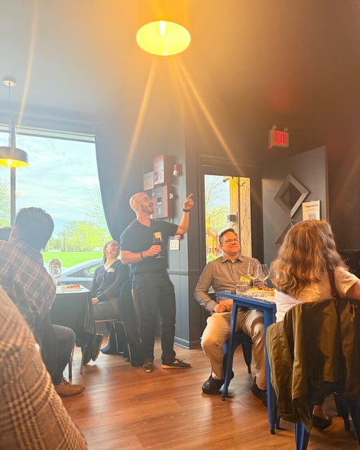 A man stands in a restaurant, holding a wine glass and pointing upwards as he speaks to a group of people seated at tables.