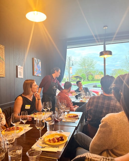 A group of people are gathered around a long wooden table in a restaurant for a wine tasting event, with a server pouring wine for a guest.