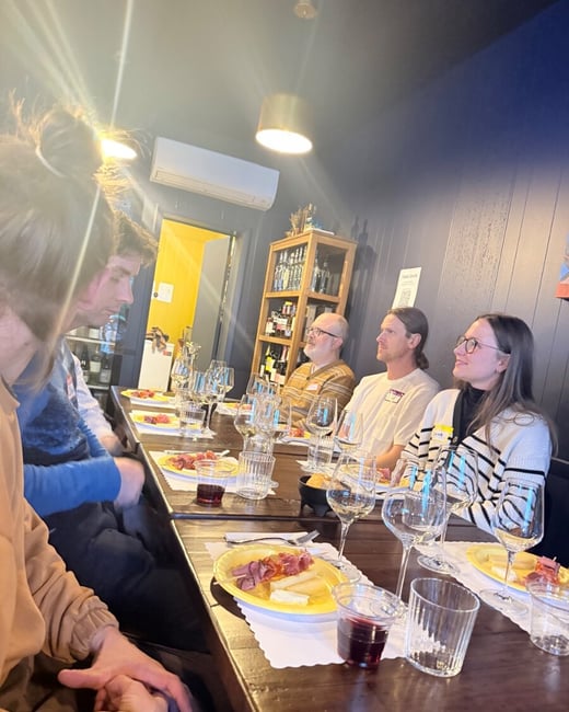 A group of people are gathered around a long wooden table in a dimly lit indoor setting, enjoying wine and small plates of food.