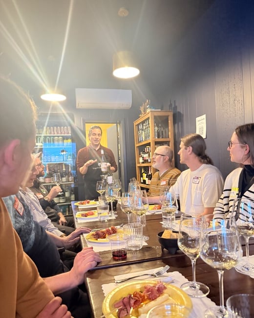 A group of people are gathered around a long wooden table for a wine tasting event, listening to a speaker presenting a bottle of wine.