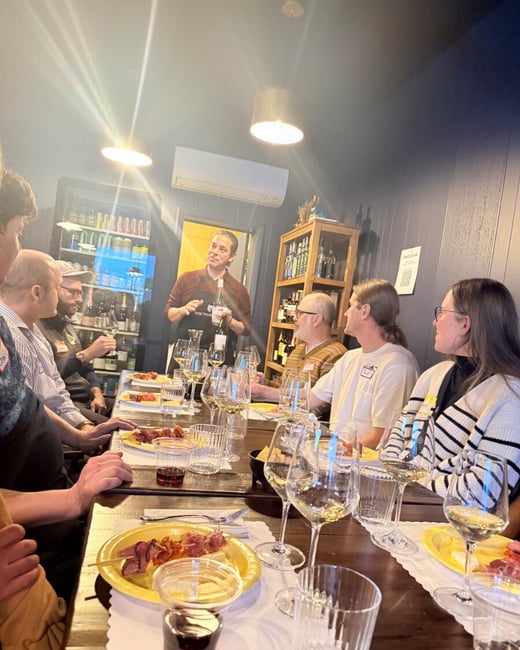 A group of people are gathered around a long wooden table for a wine tasting event, listening to a presenter standing at the head of the table.