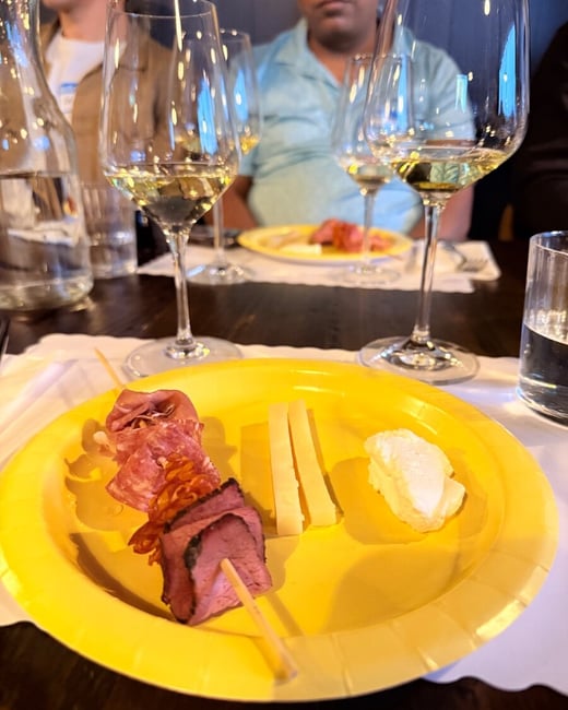 A close-up photograph of a yellow plate with meat skewers and cheese, with wine glasses and people in the background at what appears to be a social event.