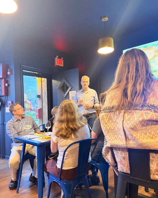 A man stands and addresses a small group of people seated at a table in a dimly lit room, likely during a wine tasting event.