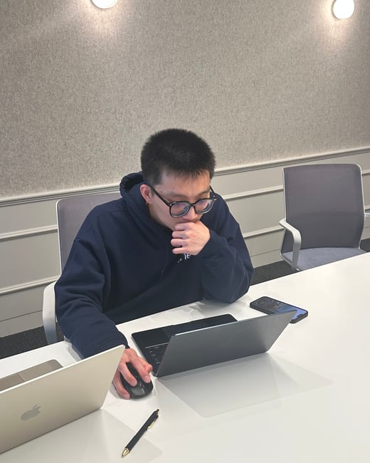 A young man wearing glasses and a navy hoodie sits at a white desk, focused intently on his laptop in an office setting.