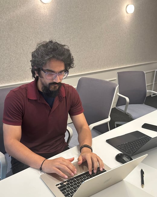 A man with curly hair and a beard is sitting at a white desk, focused on typing on a silver laptop in an office setting.