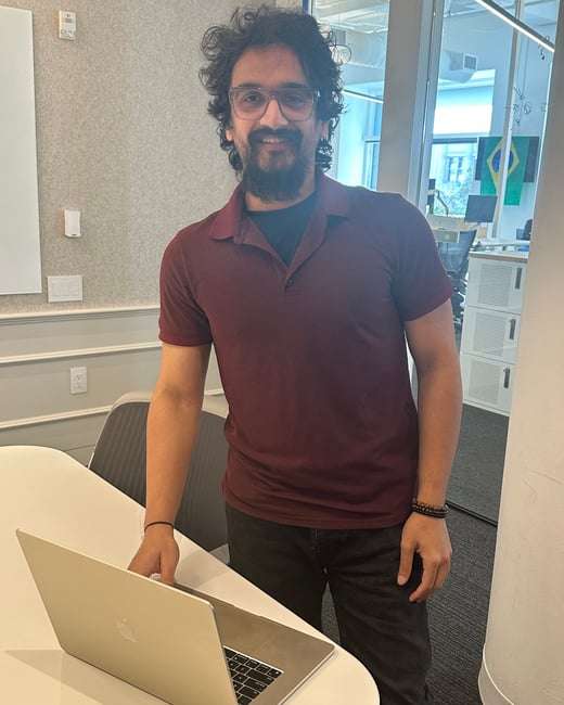 A man with a beard and glasses, wearing a maroon polo shirt, stands smiling in an office next to a laptop on a white table.