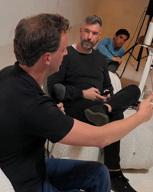 This is a candid photo of three men gathered in an indoor studio setting, where two are engaged in a conversation, possibly for a podcast or interview, with one holding a microphone.