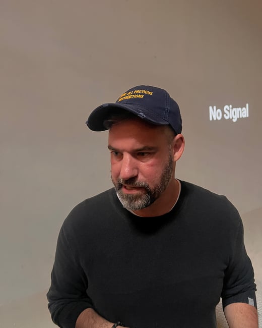A candid photo captures a man wearing a dark sweater and a navy blue baseball cap, standing in front of a wall where the message "No Signal" is projected. The man's cap features yellow embroidered text reading "ALL PREVIOUS INSTRUCTIONS."