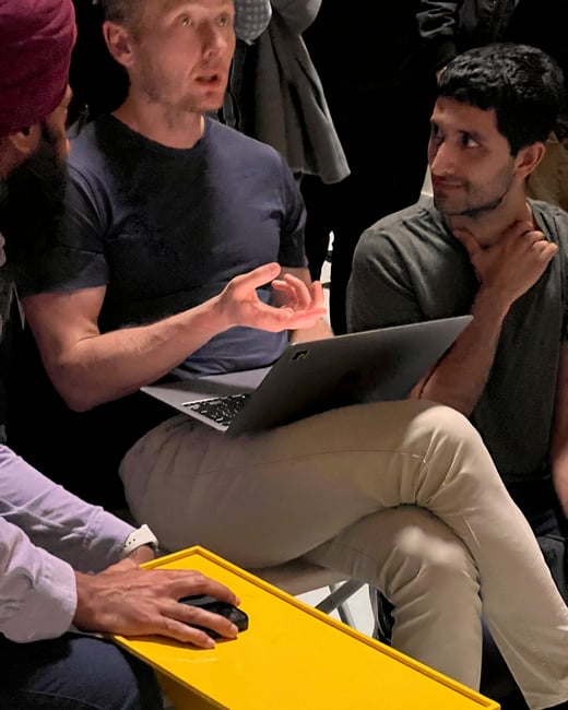 A close-up photograph captures three men gathered in a low-light setting, engaged in a discussion; one man is speaking while holding a laptop and another is using a mouse on a bright yellow surface.