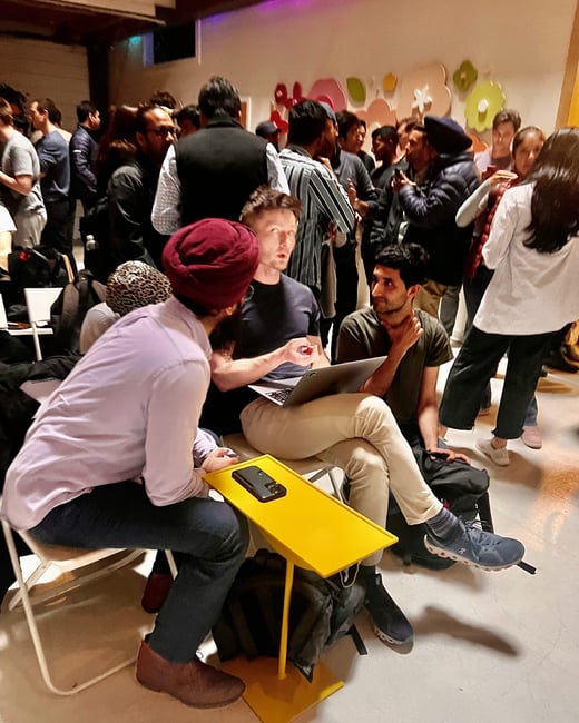 A candid photograph capturing a dense indoor social gathering or networking event, focusing on three men in the foreground engaged in a discussion around a laptop and a bright yellow side table.