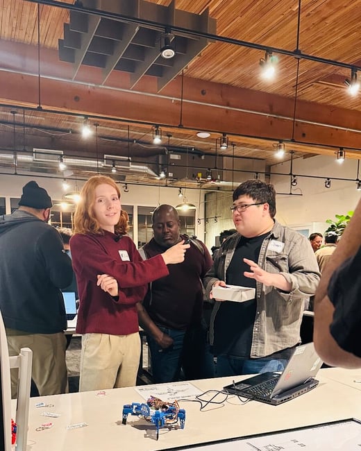 A candid photograph captures a group of people gathered around a table indoors, discussing a small blue robotic device and a framed technical diagram, likely during a project demonstration or event.