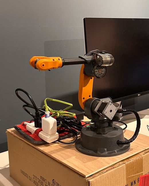 A small, articulated robotic arm, featuring orange and black components, rests on top of a cardboard shipping box along with tangled cables and a white power adapter. The scene is set indoors on a desk, with a large, dark computer monitor and a muted gray wall visible in the background.