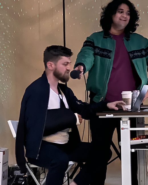 A candid indoor photograph showing two men near a table, one sitting with a black arm sling speaking intensely into a microphone, while the other stands holding the mic and smiling. The background is a light wall featuring a sparkling, low-light effect.