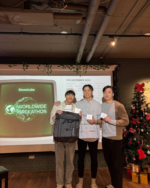 A photograph captures three young men posing and smiling in front of a projection screen displaying information about the 'ElevenLabs Worldwide Hackathon' and a decorated Christmas tree, likely at an awards ceremony.