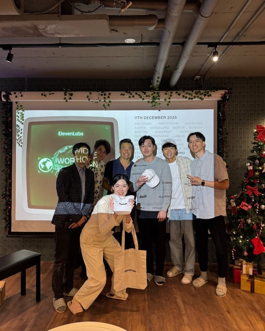 A group of seven people, likely colleagues, pose for a group photo indoors in front of a large projection screen displaying city names and the ElevenLabs logo, next to a decorated Christmas tree.