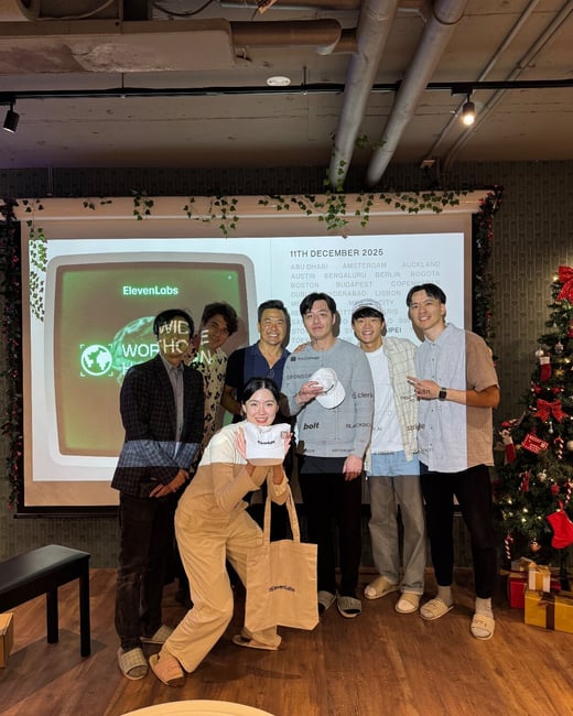 A group of seven people, primarily young adults, poses indoors in front of a projection screen displaying the ElevenLabs logo and a list of cities, next to a decorated Christmas tree. The woman in the foreground holds up a white hat and a tote bag, both featuring the ElevenLabs brand.
