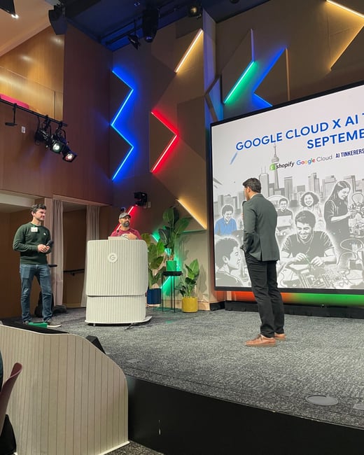 A photograph capturing an indoor corporate event or presentation, showing three people on a stage in front of a large screen displaying event details for "Google Cloud X AI" and a decorative wall illuminated by colorful LED lights.