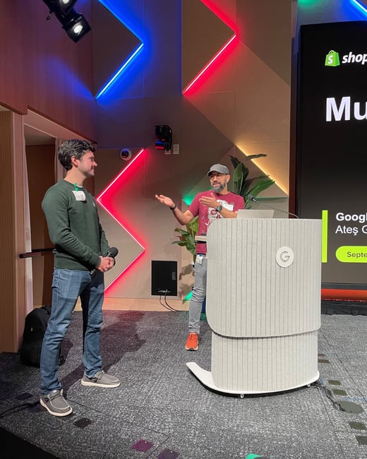 A photograph capturing two men on a stage during an event, featuring dynamic red and blue neon lighting in the background and a large screen displaying presentation content, including the Shopify logo and partial text.