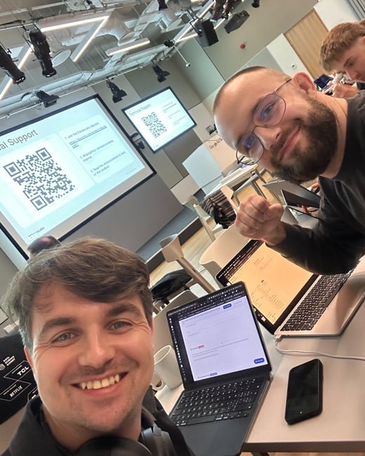 A casual, high-angle selfie taken indoors at what appears to be a tech event or workshop, showing two men smiling directly at the camera while working on laptops, with presentation screens visible in the background.