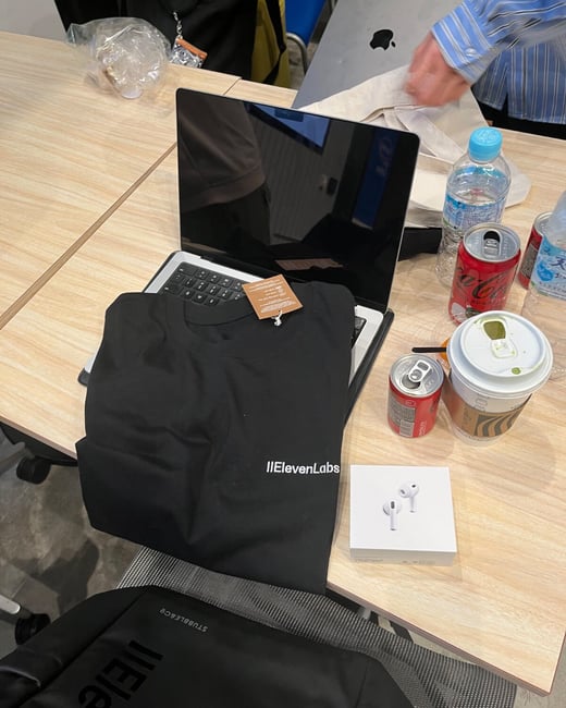 A high-angle photograph shows various items arranged on a light wooden table, including a black t-shirt branded 'IIElevenLabs,' an open laptop, an Apple AirPods box, several drink cans (Coca-Cola), and water bottles.