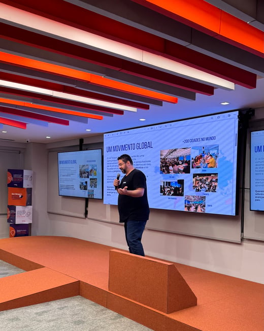 A man stands on an orange stage giving a presentation in front of a large screen displaying information about a global AI movement.