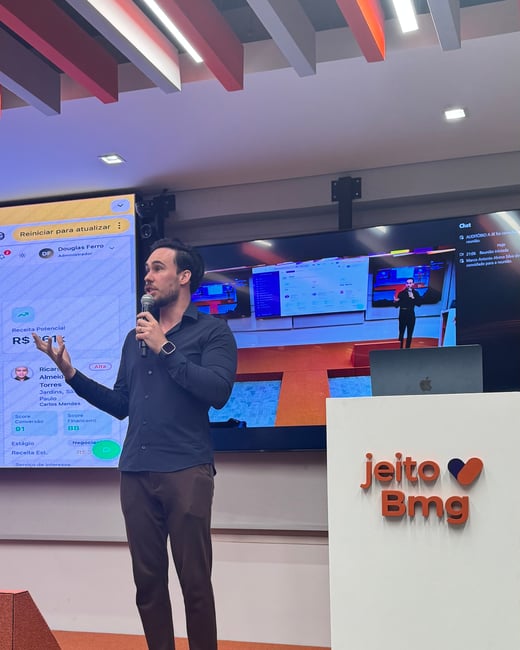 A man in a dark shirt and brown trousers is giving a presentation on a stage, holding a microphone. Behind him, large screens display data dashboards and a video feed of the event, next to a podium with the 'jeito Bmg' logo.
